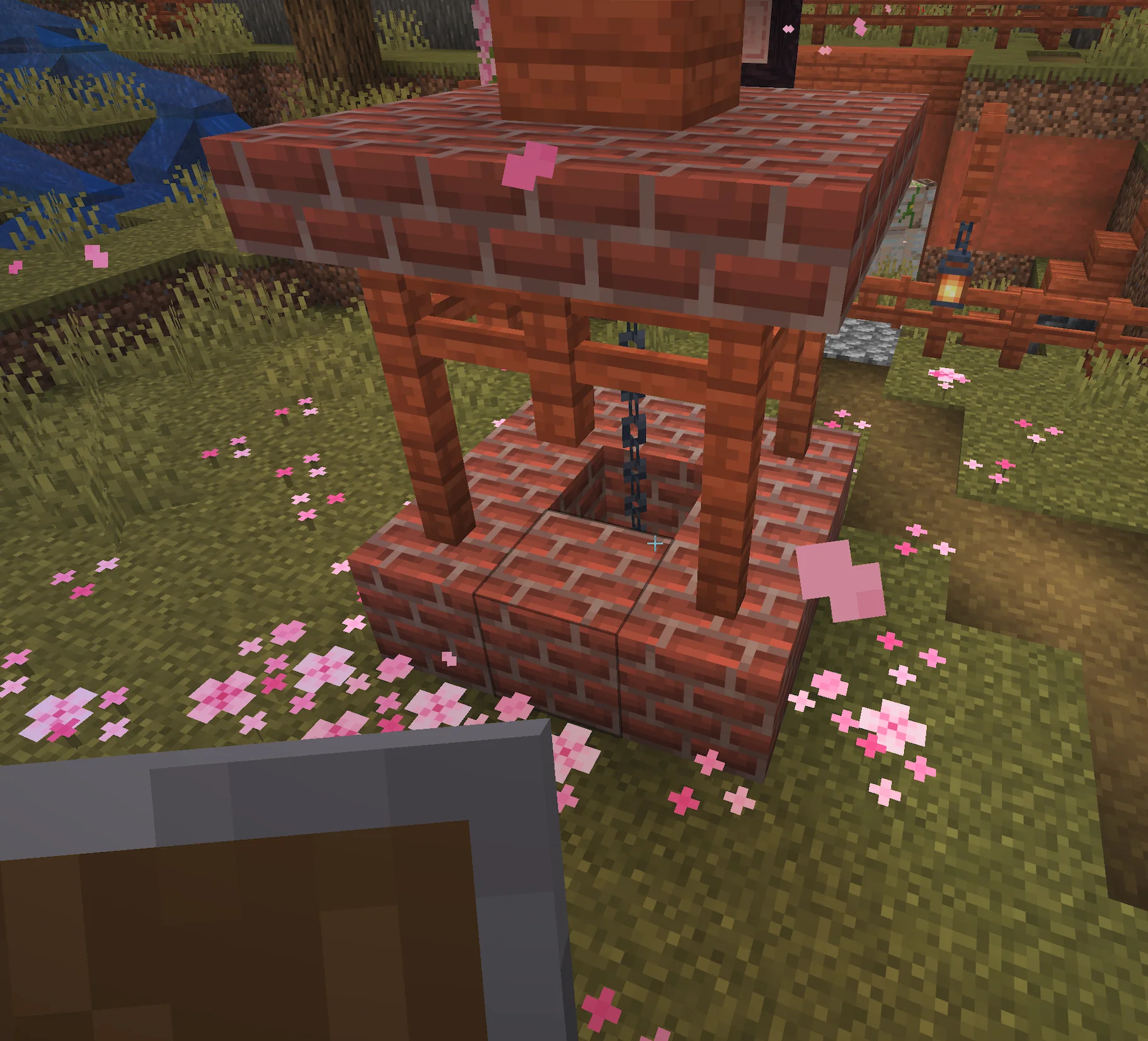 a screenshot from Minecraft of a brick well with a chain going down into it, surrounded by grass and cherry blossom petals