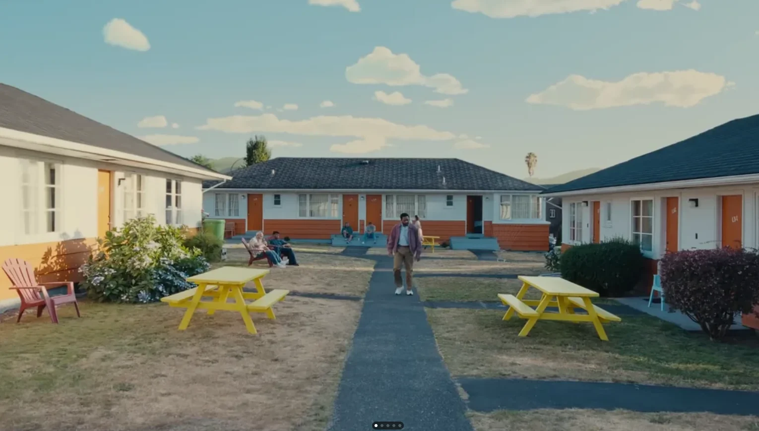 An image of the main character in the Backrooms movie walking in-between housing units down a black-top sidewalk, with two feverishly yellow picnic tables on each side of him, and a sky that looks fake
