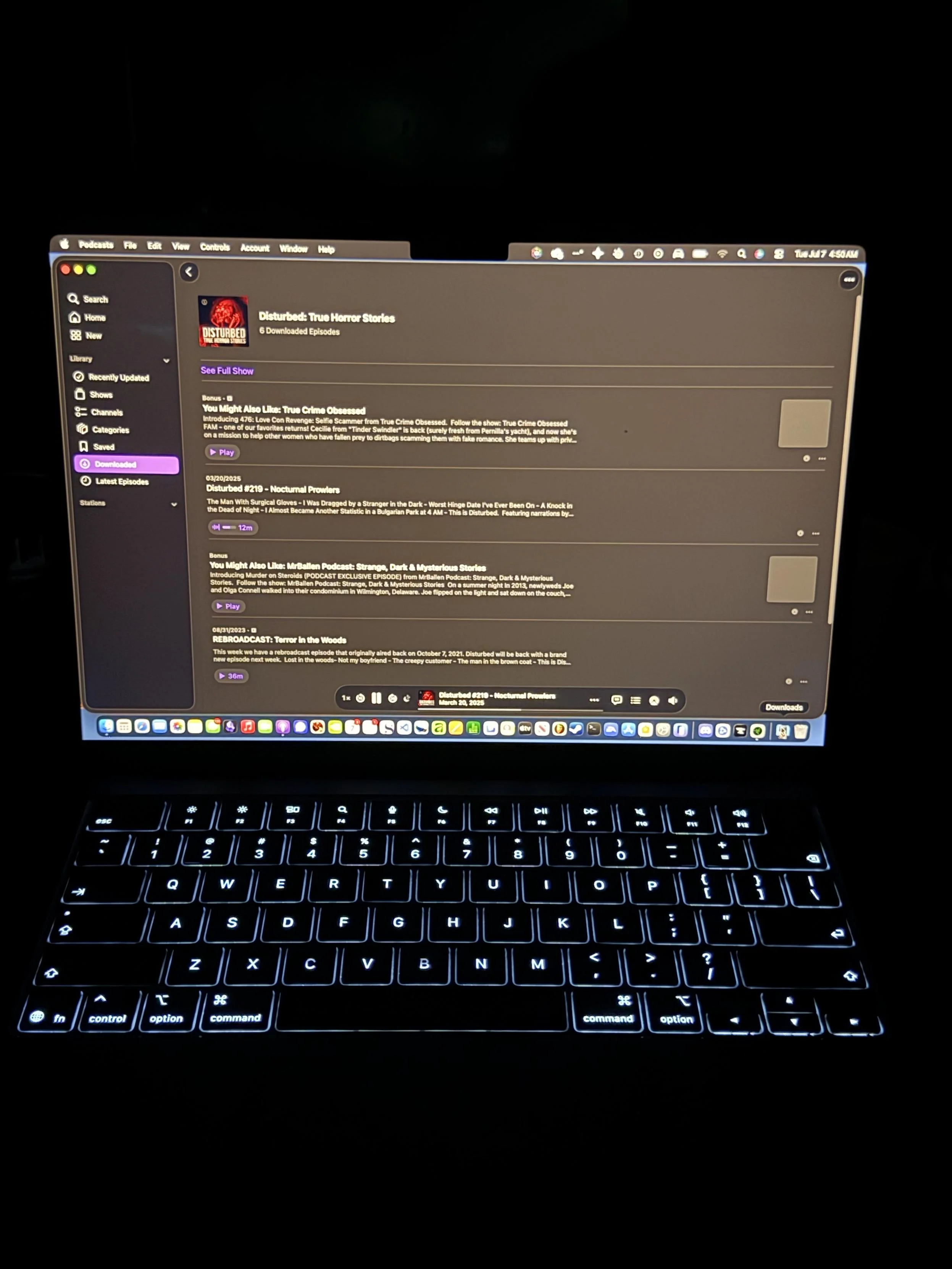 A laptop displaying a podcast app interface in dark mode. The screen shows the podcast "Disturbed: True Horror Stories," with various downloaded episodes listed, including titles and times. The laptop keyboard is illuminated.