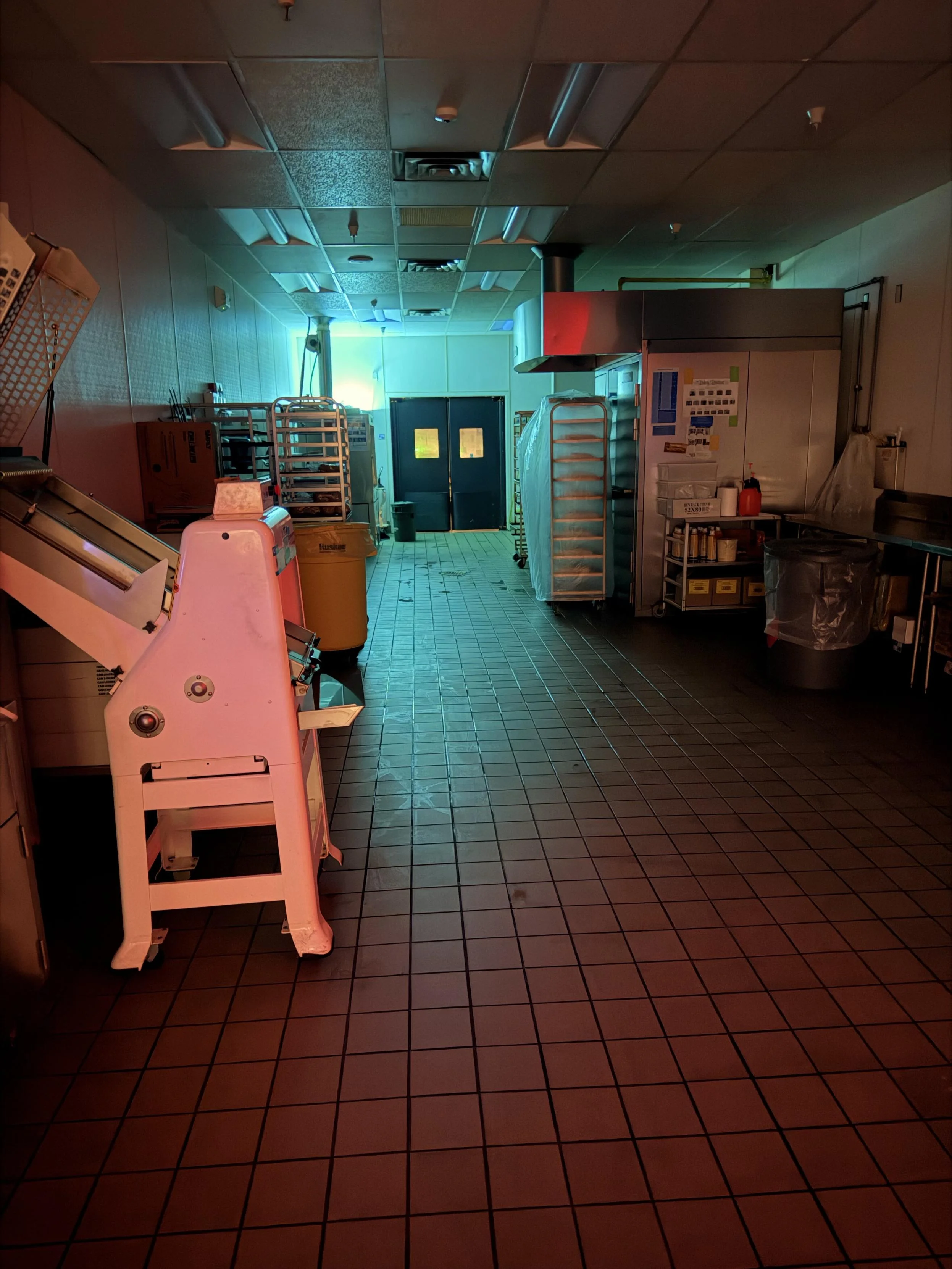 A dimly lit commercial kitchen with teal and red lighting, featuring stainless steel appliances, a large sink, racks for trays, and various kitchen equipment. The far end shows a double-door entrance. The floor is tiled, and there are shadows cast from